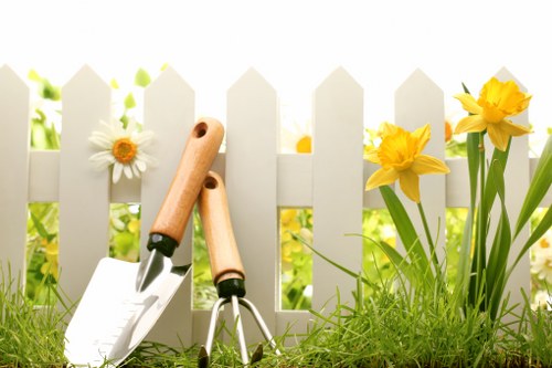 A close-up of a small garden area in Lewisham, featuring a white picket fence in the background. In the foreground, there is a patch of lush, green grass with a variety of flowers growing alongside the fence, including white daisies and bright yellow daffodils. The garden surfaces include a well-maintained lawn and nearby soil beds. Garden tools such as a hand trowel and pruning shears rest on the ground, suggesting recent or ongoing gardening work. The scene is illuminated by natural sunlight, indicating clear weather, with soft shadows cast across the grass and plants. This image captures a neat, well-kept outdoor space suitable for gardening and lawn care services provided by companies like Hedge Trimming Lewisham, showcasing typical elements of a landscaped front or back garden in the Lewisham area, with a focus on plant growth, garden maintenance, and outdoor aesthetics.