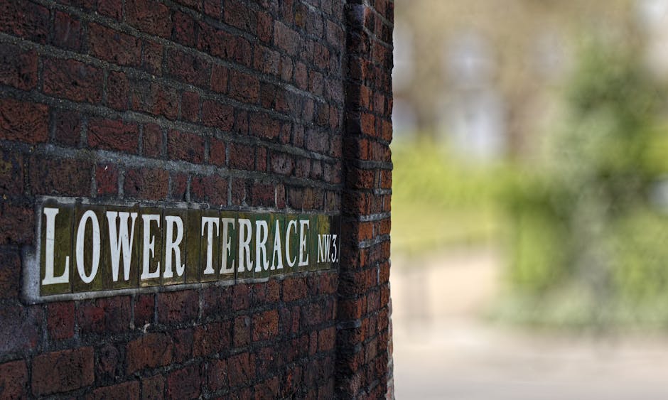 A close-up view of a brick wall with a metal street sign reading 'LOWER TERRACE NW3' attached to it. The bricks are dark red with a rough texture, and the sign features white lettering on a green back